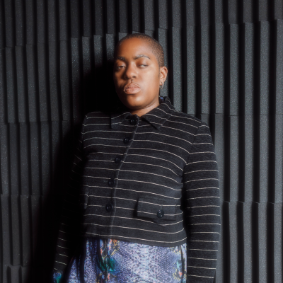 Onyeka Igwe, wearing a black jacket and standing against a textured black background