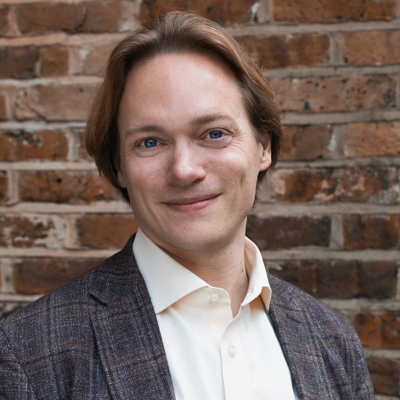 Tom Littler, who has mid-length red/brown hair and is wearing a grey suit with pale shirt