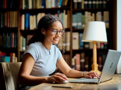 A female students sits in the library working at a laptop.