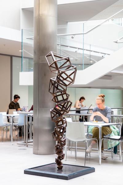 Conrad Shawcross’s Paradigm Chamfer (Structural) (2015), a tall bronze sculpture composed of interlocking geometric frames, displayed in the atrium of Oxford’s Mathematical Institute. Students are seated at tables nearby, working and eating in the bright, modern space.