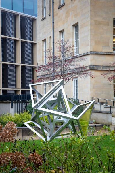 Plosion 1 (Yellow) Large (2013) by Conrad Shawcross, an outdoor sculpture at Oxford’s Mathematical Institute. The aluminium structure features interlocking geometric frames in a dynamic, partially exploded form, positioned on a lawn with surrounding plants and modern institutional buildings in the background.