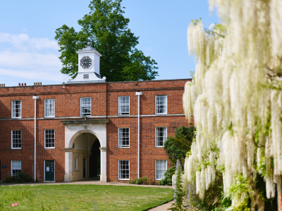 LMH’s red-brick clock tower rises above a historic college building and arched entranceway, framed by mature trees and flowering wisteria. Sunlight illuminates the lawns and gardens in the foreground, highlighting the beauty of the College grounds.