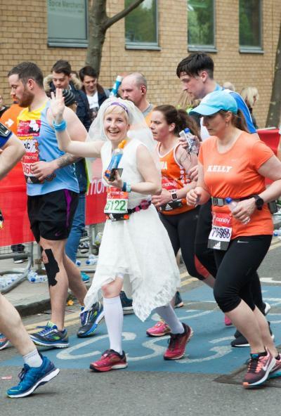 Jackie Scully running the London Marathon in a wedding dress and veil, smiling and waving at the crowd while holding a drink, surrounded by other runners.