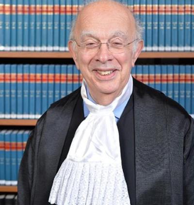 A man with greying hair wearing a Judge's robe sits in front of a shelf of blue spined law books