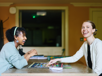 Two students sit across from each other at a table, laughing as they play a board game together. The scene captures a relaxed and friendly atmosphere, with game pieces spread across the table and warm light filling the room.