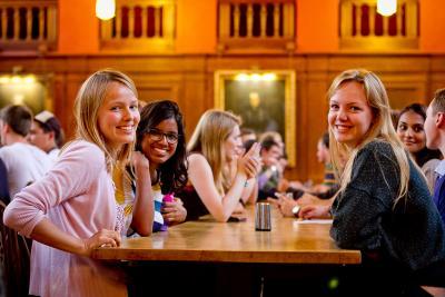 A group fo female students smile to the camera while sitting around a dining hall table.