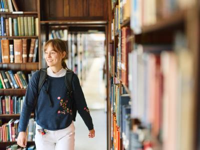 A student walks through the library between tall bookshelves, carrying a backpack and smiling. Warm natural light and rows of books create a welcoming study environment.