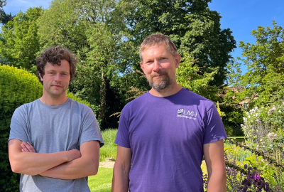 Two men in casual t-shirts standing in some gardens with blue sky and trees in the background