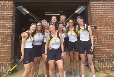 Nine women wearing blue and white lycra rowing suits standing outside a red brick boathouse building