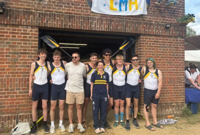 Group of nine men wearing blue and white lycra rowing outfits, standing outside of a red brick boathouse building. The words LMH can be seen written in bright blue and yellow on a banner hanging down from above.