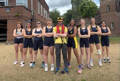 A group of eight women, and one man, stand in a v formation, with arms crossed, on the banks of the river. They wear dark navy lycra rowing suits.