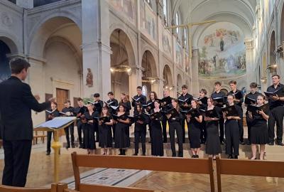 The Lady Margaret Hall Choir performing in Notre-Dame-des-Champs, Paris, conducted by a director at the front. The singers are dressed in black and standing inside the ornate church.