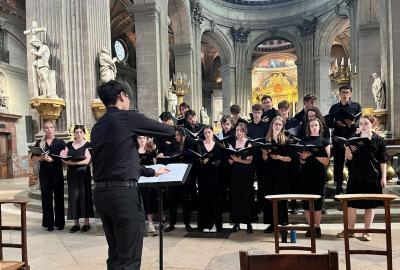 The Lady Margaret Hall Choir performing in Saint-Sulpice, Paris. The choir, dressed in black, stands in front of ornate columns and statues, with a conductor leading them from the front. Golden candelabras and a brightly decorated altar can be seen in the background.