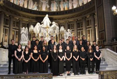 The Lady Margaret Hall Choir inside L’église de la Madeleine, Paris, posed on the altar steps in front of statues and ornate gold candelabras. The choir is dressed in black, standing beneath the church’s painted dome and decorative columns.