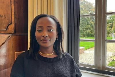 Dr. Makena Micheni sitting indoors by a large window with a garden view, wearing a black sweater and gold necklace, smiling softly at the camera