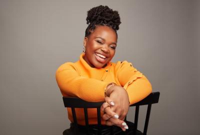 Dr Vee Kativhu sits on a backwards chair smiling expressively at the camera