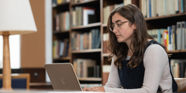 A woman with long dark brown hair and glasses sits in a library working on her computer